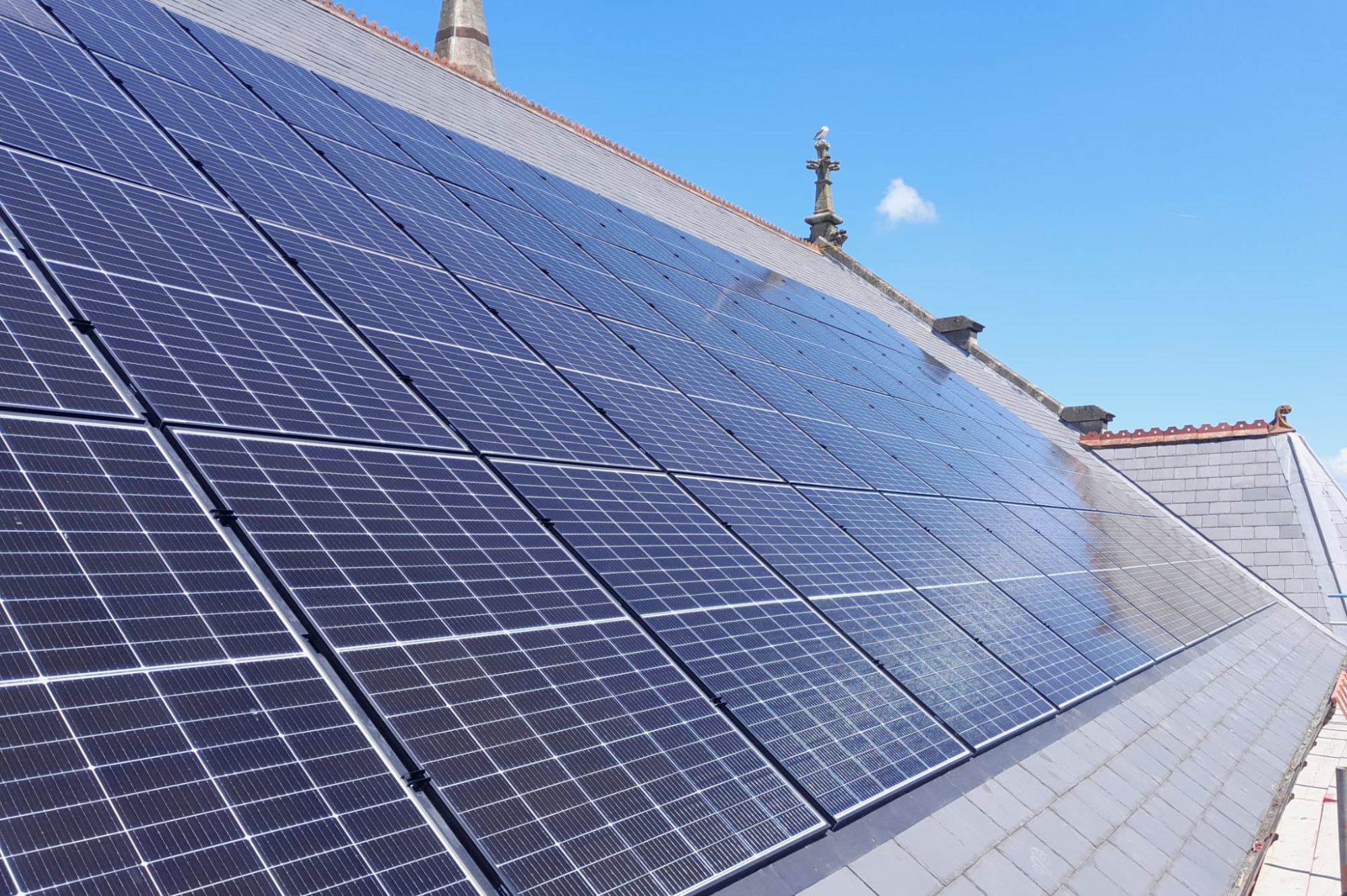 Solar panels on the side of a sloping church roof with a seagull perched on a stone cross in the background.