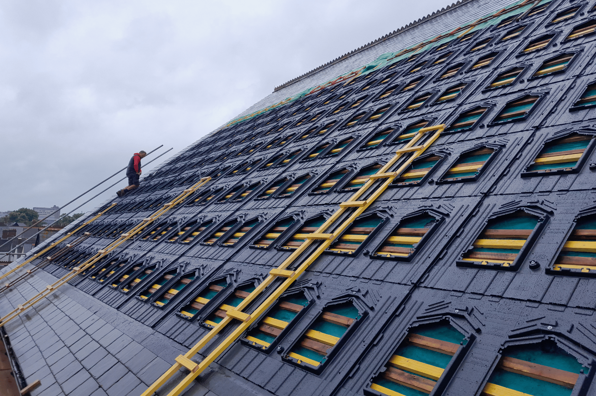 Fittings for in-roof solar panels on the roof of The Gate Arts Centre, punctuated by yellow ladders, with an InspireGreen installer kneeling in the background.